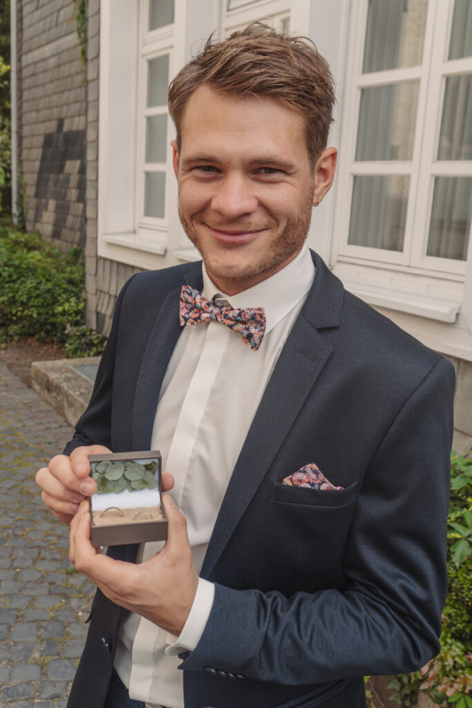 Bräutigam im Anzug hält eine Ringbox mit Eheringen in der Hand.