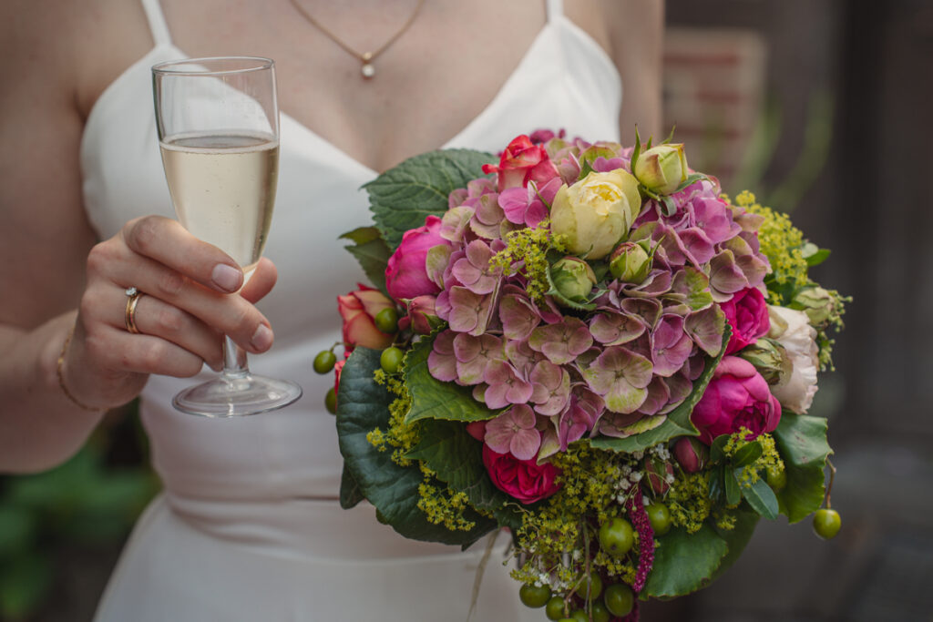 Braut hält ihren farbenfrohen Brautstrauß und ein Glas Sekt in der Hand.