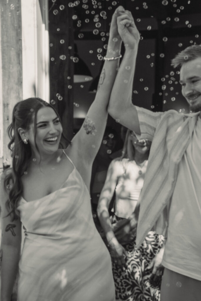 Brautpaar hebt jubelnd die Hände beim Auszug nach der Trauung, umgeben von Seifenblasen.