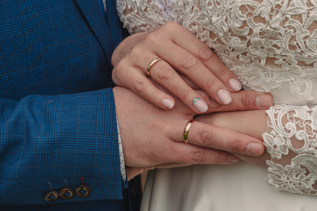 Detailaufnahme der Hände des Brautpaares mit goldenen Eheringen vor dem Brautkleid.