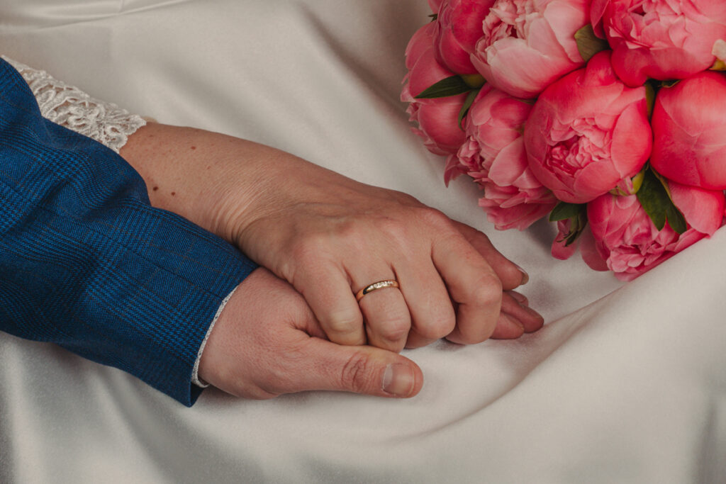 Detailaufnahme der ineinander verschränkten Hände eines Brautpaares mit Eheringen und pinkem Brautstrauß.