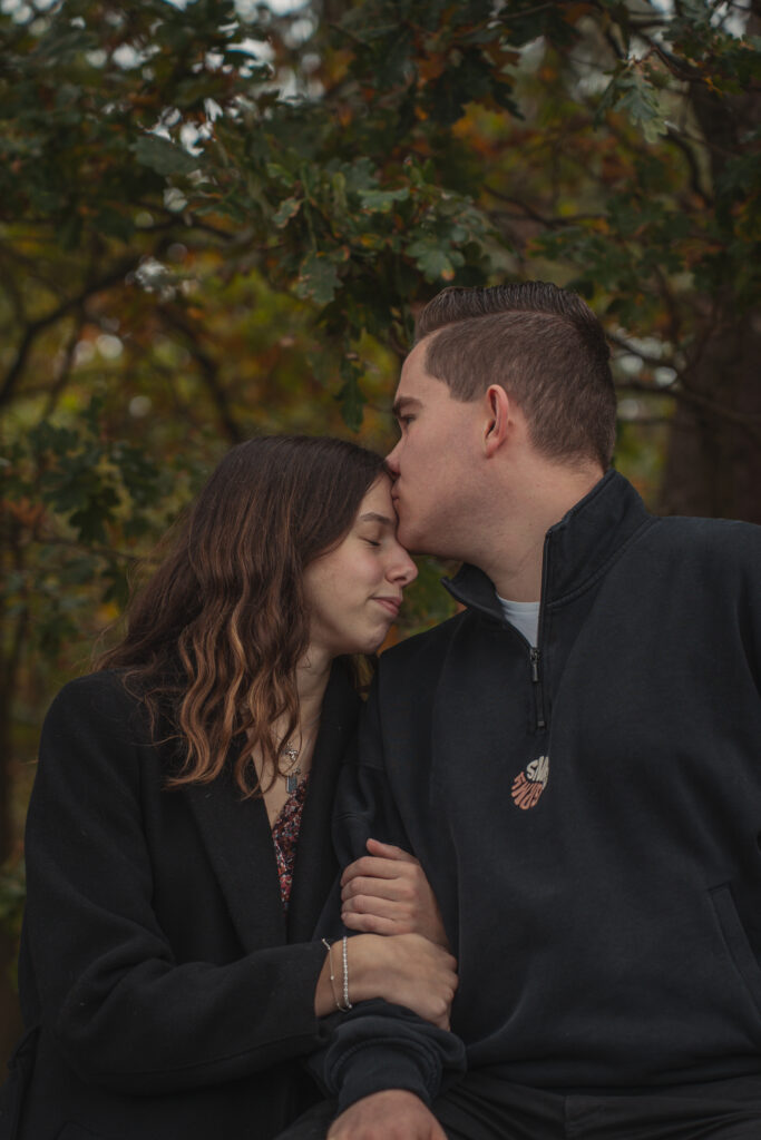 Er küsst sie zärtlich auf die Stirn unter herbstlichen Bäumen beim Paarshooting.