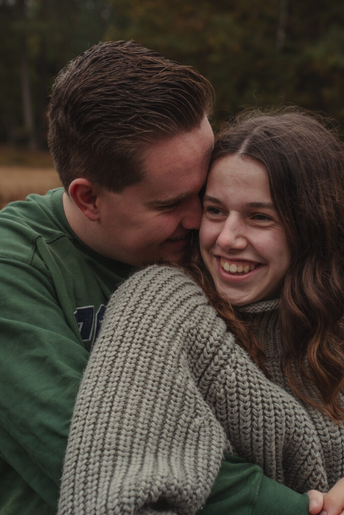 Er umarmt sie von hinten, beide lachen vertraut beim Paarshooting im Freien.