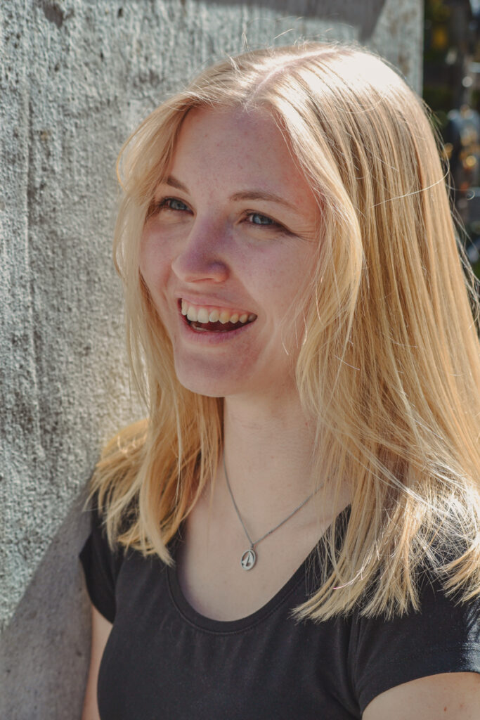 Einzelportrait einer lächelnden Frau im natürlichen Licht vor einer Betonwand.