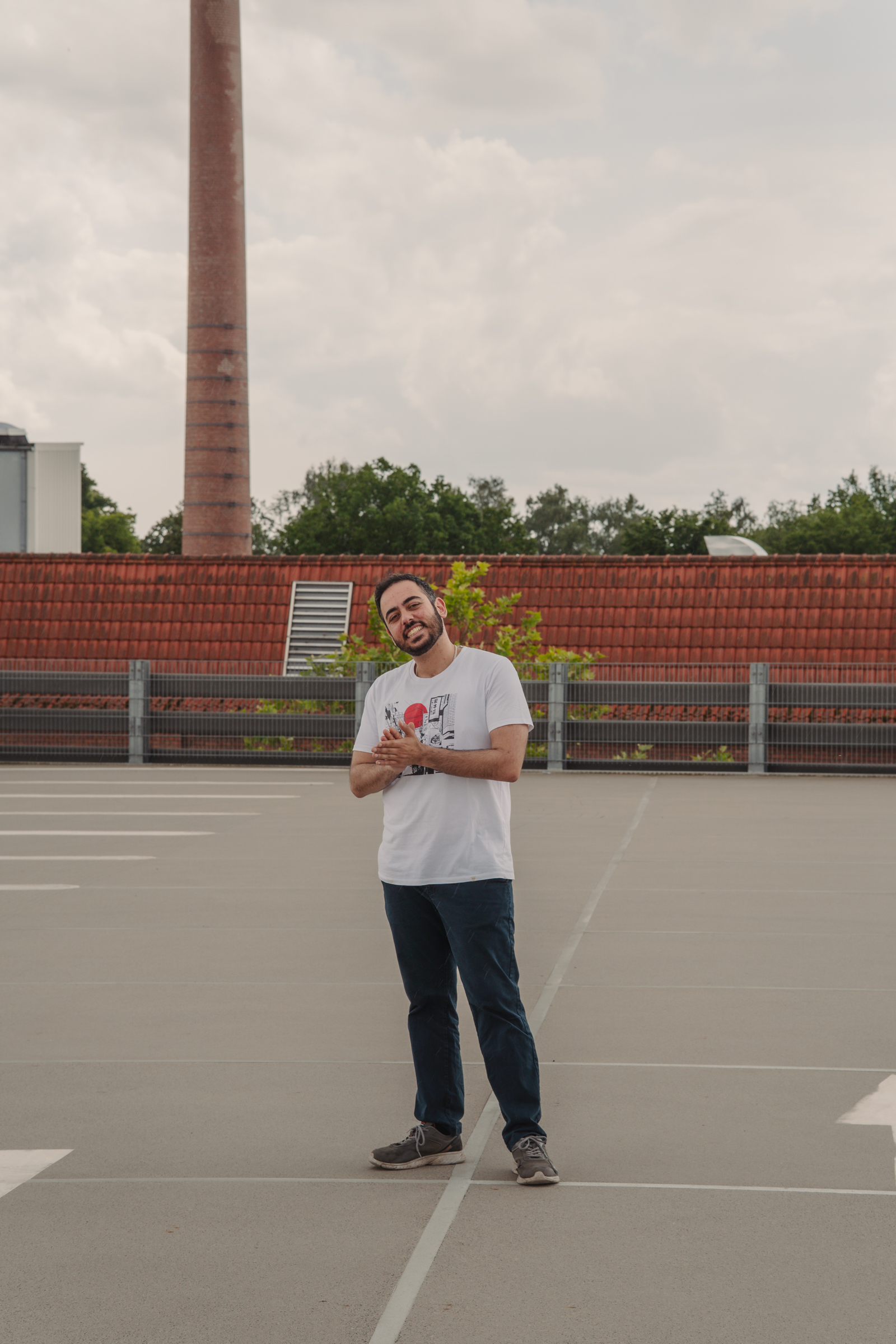 Ganzkörperportrait eines Mannes auf einem Parkdeck mit industrieller Kulisse im Hintergrund.
