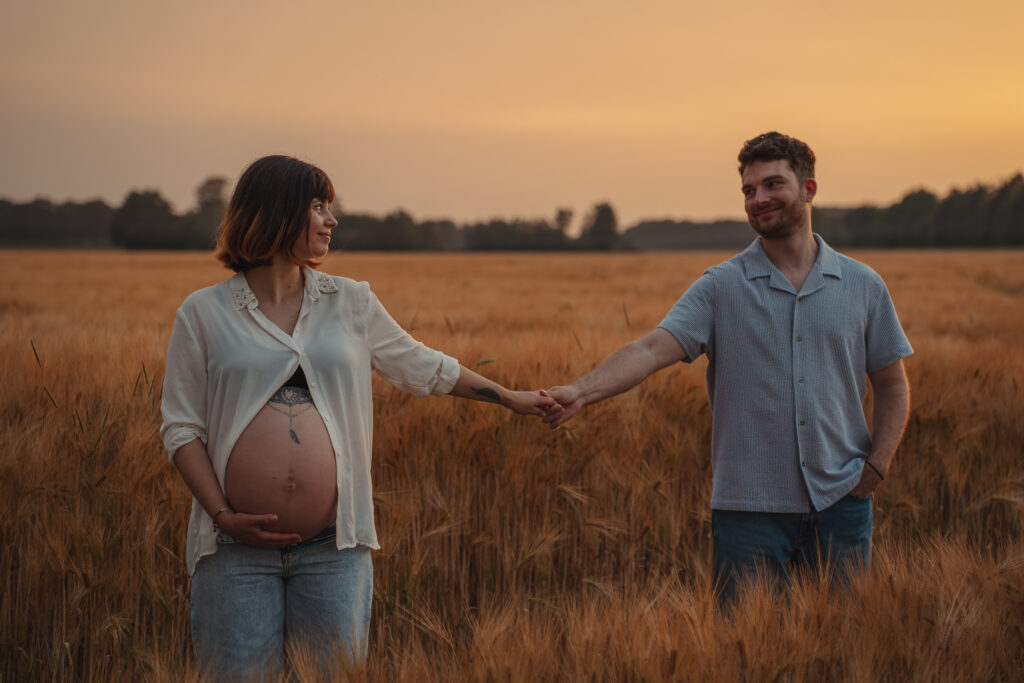 Werdende Eltern halten sich im Kornfeld an den Händen und freuen sich auf ihr Baby.