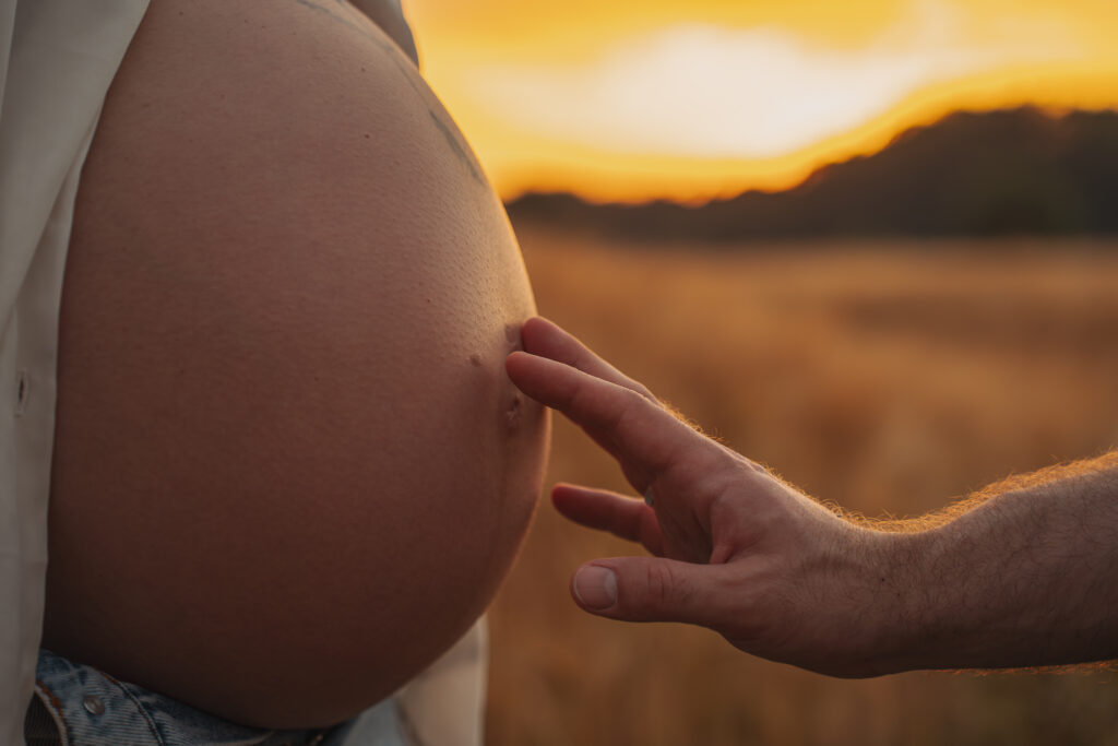Die Hand des werdenden Papas berührt liebevoll den Babybauch seiner Partnerin im warmen Abendlicht.