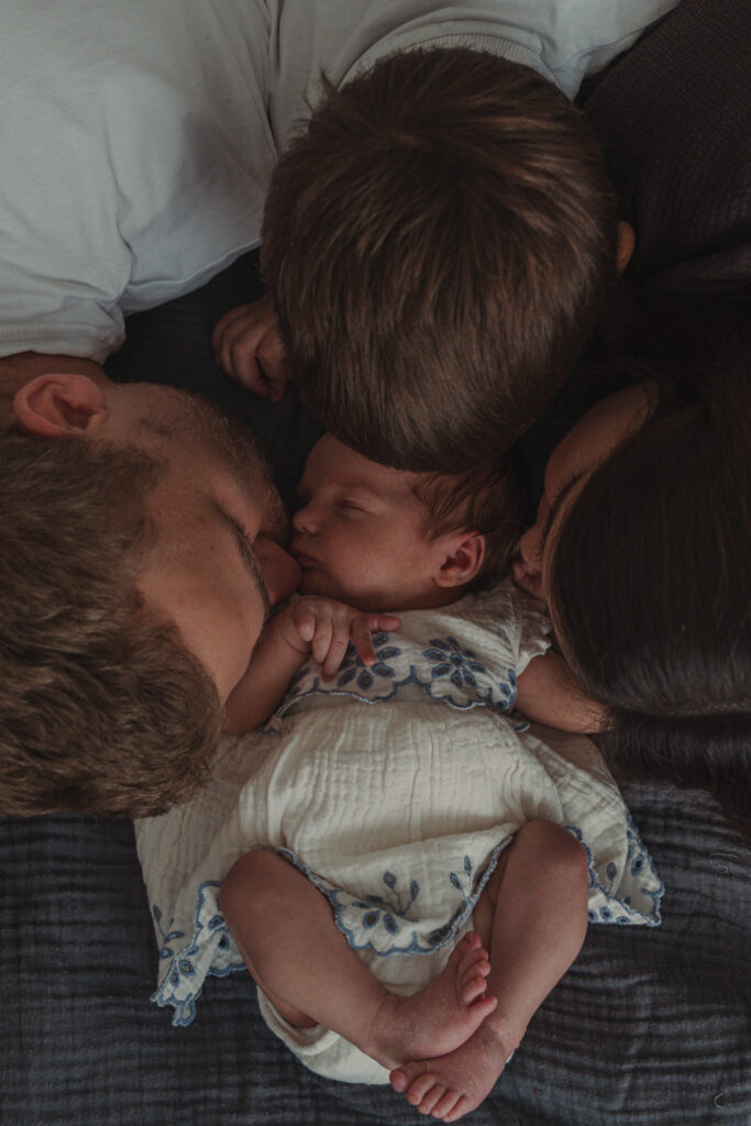 Familie kuschelt gemeinsam um ihr schlafendes Neugeborenes auf dem Bett.